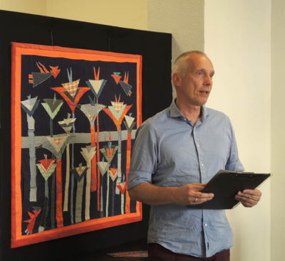 Eröffnungsredner Alexander Lange, im Hintergrund  die Textilcollage »Neugierde-beidseitig« von 2012